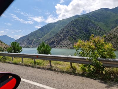 A scenic view captures a highway running alongside a calm, turquoise lake in Ardanuç, Turkey. The road, with a white line marking its edge, is visible in the foreground, with a portion of a red car's side mirror reflecting the scene. A metal guardrail separates the road from the lake. On the left side of the lake, two evergreen trees stand tall, while lush green vegetation grows on the mountainous terrain in the background. The sky is a clear blue with scattered white clouds, and power lines stretch across the frame.