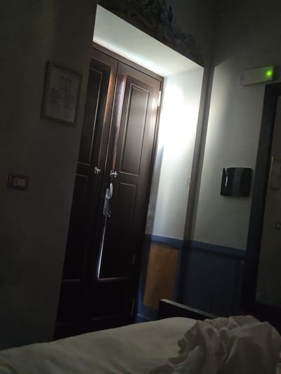 The image depicts an interior scene, likely a hotel room, in Pozzuoli, Italy. Dominating the left side of the frame is a large, dark wooden double door with ornate handles. A sliver of bright light emanates from the crack between the doors, suggesting an open space beyond. To the left of the door, a framed document is affixed to a pale gray wall. Below the document, a light switch plate is visible.
The right side of the image shows a white wall with a blue and orange horizontal stripe pattern near the bottom. A black, rectangular object, possibly a ventilation grille or a decorative panel, is mounted on the wall. Further to the right, a green emergency exit light is illuminated. The foreground of the image is occupied by a rumpled white sheet, suggesting an unmade bed. The lighting is dim, with significant shadows cast across the scene. The ceiling features a decorative mural, partially obscured by darkness.