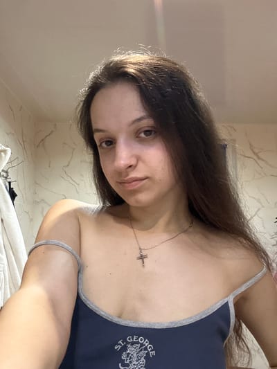 A young woman with long, dark brown hair is captured in a close-up, selfie-style shot. She is wearing a dark blue tank top with gray trim and a silver cross necklace. The background appears to be a bathroom, with marble-patterned tiles on the wall and a towel rack with towels visible on the left. The lighting suggests an indoor setting, possibly illuminated by artificial light. The image focuses on the woman's face, shoulders, and upper chest, with a slight tilt of her head and a neutral expression. There is no specific indication of the location being Postoyata, Ukraine.