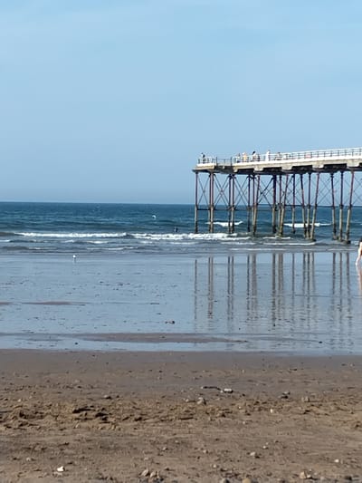 This is a daytime, eye-level, outdoor medium shot of the Saltburn Pier in Saltburn by the Sea, United Kingdom. The pier extends out over the ocean, supported by numerous metal stilts. People are visible walking on the pier. The ocean is a dark blue with white waves. The tide is low, with a wet sand beach in the foreground, reflecting the pier and the sky. There are a few seagulls on the wet sand. The sky is clear and blue.