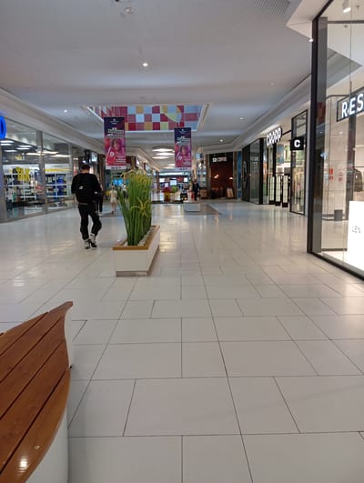 The image shows the interior of a shopping mall with bright white tiled floors and a high, white, corrugated ceiling. A man in black pants and a black shirt, wearing roller skates, is walking away from the camera. He is carrying a black backpack. To his left, a rectangular planter filled with tall, green artificial plants sits on the floor. In the background, several shopfronts are visible. One sign reads "RE." Another sign has the word "CROPP" in large letters. Further down the corridor, a shop called "SOI COFFEE" is advertised. To the left, banners hang from the ceiling, one with the text "TV NAGRADZA!" The overall impression is that of a modern, well-lit shopping environment. The location is the CAMPORE Shopping Centre in Wołomin, Poland.