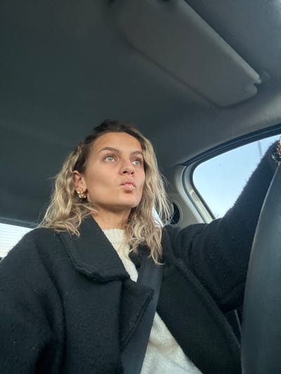 A woman sits in the driver's seat of a car. She is wearing a black coat over a white sweater, her blonde curly hair is framing her face, and she has on decorative earrings. The sun visor is visible above her. She is wearing a seatbelt. The car's ceiling is visible. The woman is looking upwards and slightly to the left with a thoughtful expression. The scene is well-lit with natural light from the car windows.