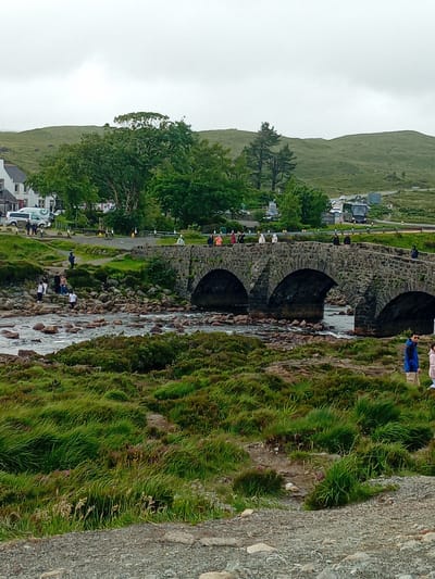 An overcast day frames a stone arched bridge spanning a shallow, rocky river on the Isle of Skye, United Kingdom. The bridge, constructed with rough-hewn stone, features three arches, with the central one being the largest. The riverbed is strewn with numerous rocks and boulders of varying sizes, with clear water flowing between them. Lush green vegetation, including grass and heather, covers the riverbanks and the surrounding hills.
Several people are visible in the scene. On the bridge, individuals are walking and standing, observing the surroundings. Near the river's edge, a group of people, including adults and children, are gathered, some appearing to interact with the water or the rocks. In the foreground, two individuals stand facing away from the camera, one wearing a blue jacket and the other a light-colored outfit.
In the background, a cluster of buildings, including white structures with dark roofs, line a road. Vehicles, including a white van and a dark bus, are parked or driving along this road. The landscape beyond the buildings consists of gently rolling green hills, indicating a rural or semi-rural setting. The overall atmosphere is one of natural beauty and leisure activity.