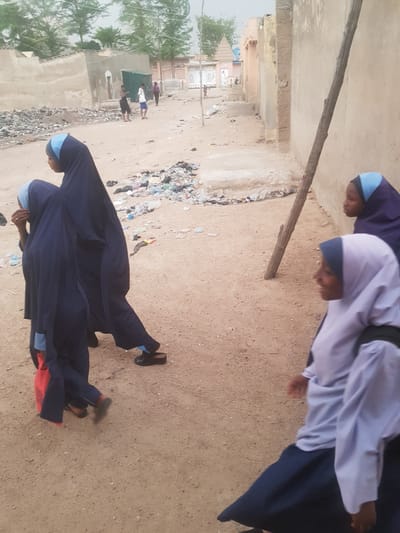 The image shows a street scene in Dutsinma A, Nigeria. In the foreground, two people wearing navy blue robes and light blue head coverings are walking on a sandy surface. The person in the front also has a red object near their feet. Further back, other people are walking on a dirt road, with buildings and trees visible in the background. There is also trash and debris scattered along the road.
