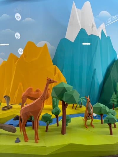 This image captures a static, multi-layered diorama display located in Guangming District, China. The scene is rendered in a low-polygon geometric style, presenting a landscape with diverse ecosystems and animal figures.

The foreground consists of undulating green terrain, bisected by a winding blue river. A distinct light brown, sandy area occupies the left side of the foreground. Various geometric trees with green polygonal canopies and brown trunks are interspersed throughout the green areas; one palm tree is also present.

Animal figures include an orange, low-poly giraffe standing upright in the center-left, and an orange, two-humped camel positioned behind the giraffe. On the sandy ground to the far left, a coiled brown cobra is visible. To the right, an orange, low-poly kangaroo with a joey in its pouch stands. A dark-colored, low-poly quadruped is partially obscured near the river on the left. Several green columnar cacti are present within the sandy section.

The background features prominent, stylized mountain ranges: a multi-peaked orange range on the left and a taller, multi-peaked teal range with white snow-capped peaks on the right. The sky is light blue with stylized white clouds. Several circular, illuminated icons displaying animal silhouettes or symbols are visible in the sky, suggesting projected imagery or a digital screen integration. Chinese characters appear as text on the far left of the sky area and possibly on the teal mountain face. The overall display is a stationary, educational, or public art installation. No human figures are present.