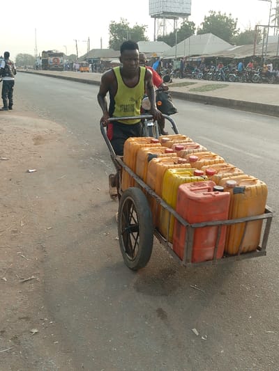 A man pulls a cart filled with yellow and orange jerrycans along a paved road. He is wearing a yellow tank top and dark shorts. Another man on a motorbike follows closely behind. In the background, a line of parked motorcycles and buildings with corrugated metal roofs are visible, along with a tall water tower. A few other people are seen in the distance.