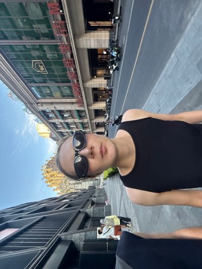 The image presents an inverted selfie taken by a person standing on a city street, looking down. The main subject, a person wearing a black tank top and sunglasses, occupies the foreground, with their head positioned at the bottom of the frame due to the upside-down orientation.

The background depicts a bustling urban street scene. On the left side of the frame (which would be the right side if the image were rotated correctly), a multi-story building with classical architectural features is prominent. Large red Chinese characters, clearly reading "中国银行" (Bank of China), are visible on its facade, alongside a distinctive corporate logo featuring a horse's head within a shield. Numerous motorcycles are parked along the sidewalk in front of this building.

On the opposite side of the street, a modern, dark-colored high-rise building with a sleek, angular design stretches upward. Further in the distance and reflected in some windows, a unique golden, ornate building with a complex, spired roof structure stands out against a clear blue sky dotted with scattered white clouds. Other pedestrians can be seen walking on the sidewalks on both sides of the street, some carrying bags. The scene is illuminated by bright daylight, suggesting a sunny day. The setting is an active urban environment, located in Putuo District, China.