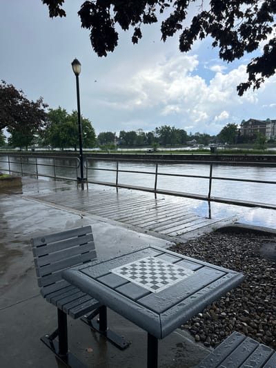 A wet, grey, overcast day in Sainte-Anne-de-Bellevue, Canada. A chessboard and bench sit on a wet concrete and wood boardwalk next to a large body of water, possibly the Ottawa River or Lac des Deux Montagnes. Trees line the far bank, with buildings visible in the distance. The chess table is set up with a standard chessboard pattern, and both the table and bench are wet from rain. A streetlamp stands nearby. The scene suggests a public park or waterfront area designed for recreation, but the weather has made it temporarily unusable.
