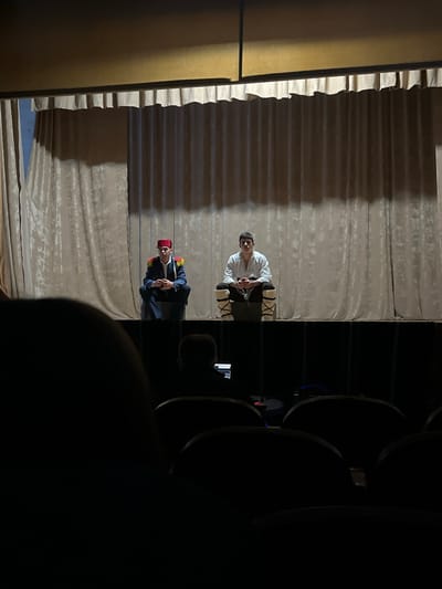 The image captures a theatrical performance viewed from the audience. A beige curtain serves as the backdrop. Two actors are on stage. One actor, wearing a white outfit and leg wrappings, sits on a chair. Chains appear to bind his hands. The other actor stands, wearing a dark blue uniform with gold trim and a red fez. He holds a microphone and seems to be speaking. Seating is visible in the foreground and to the right, suggesting an auditorium setting. The location appears to be a performance in Voiniagovo, Bulgaria.
