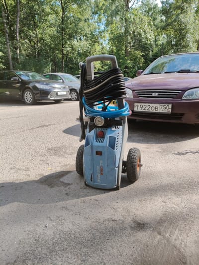 An outdoor daytime shot captures a blue pressure washer, identified by the label "Kranzle 2160TS" on its side, parked on an asphalt surface. The pressure washer has two wheels and a hose coiled on top. In the background, two cars are visible: a dark sedan on the left and a maroon Kia on the right, which displays a license plate reading "FT922OB 750 RUS". Behind the cars, a dense line of trees forms a backdrop against a bright sky. The scene appears to be in a residential area or parking lot within the city of Alfimovo, Russia.