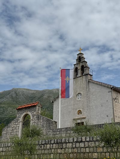 Here's a detailed description of the image:
The image depicts a building with light gray stone construction, likely a church or historical structure. The building features a multi-tiered bell tower topped with a golden cross. A flag with horizontal stripes of white, blue, and red with a centered cross is flying from a pole attached to the building.
The background includes a steep, rocky mountain with green vegetation. The sky is filled with white, puffy clouds. A stone wall with vegetation growing on it is visible on the right side of the frame, and a small arched structure with a red-tiled roof appears in the lower part of the frame.