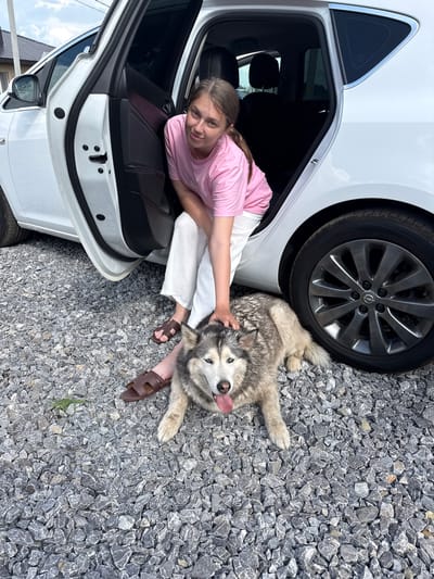 In Нагаево, Russia, a young woman with brown hair is sitting on the ground next to an open car door. She is wearing a pink t-shirt and white pants, with brown sandals on her feet. A husky dog is lying down on the gravel ground next to her, and she is petting it. The car is white, and its doors are open. The ground is covered in gray gravel, and there are some green blades of grass visible. The sky is cloudy.