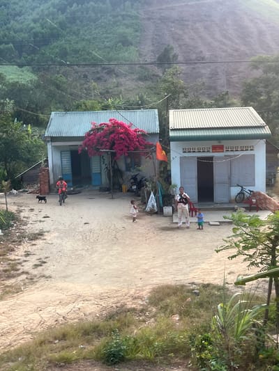 The scene depicts two single-story buildings in what appears to be a rural area of Myui, Vietnam. The building to the left is partially obscured by a large, flowering bougainvillea bush. The building to the right has an open doorway, and two adults and two children are standing in front of it. A woman on a bicycle, wearing an orange shirt, is riding on a dirt path in front of the other building. There is a dog running beside the bicycle. A motorcycle is parked in front of the building with the bougainvillea bush. In the background, there is a steep, green hillside covered in trees. The overall environment seems warm and sunny. Power lines run across the image.