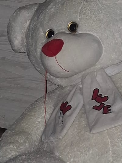 A close-up, indoor, eye-level shot shows a large, white, plush teddy bear. The bear has a large red nose and dark eyes with gold rims. It is wearing a white scarf with red heart shapes and the word "LOVE" inscribed in red letters. A thin red string hangs from the bear's mouth. The background consists of a gray, textured wall. The scene is not located in the city of Grivitsa, Bulgaria; it is an indoor setting.