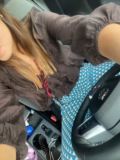 The image depicts a partial view of an individual seated in the interior of a vehicle, consistent with the driver or front passenger seat, in what is contextually specified as Carnaxide, Portugal. The subject, a female, has fair skin, visible from the lower left portion of her face with pink-tinted lips. Her long, straight hair, which is light brown to blonde, extends across her chest. She wears a dark brown, long-sleeved, collared shirt made of a textured or ruffled fabric, with elasticized cuffs. Below the shirt, a garment with a repetitive geometric pattern of blue and white triangles is visible. Around her neck or draped over her chest is a red and black striped lanyard with a silver-colored clip, to which a small red tag is attached.

The vehicle's interior features a grey upholstered seat on the left. To the right, a black steering wheel with silver accents and control buttons is partially visible. The center console includes cup holders, one containing a blue Nivea tin. Other items on the console include a black comb, a white charging cable connected to a port, and several black buttons and switches. A red seatbelt buckle is also visible. The overall scene is well-lit, suggesting daytime conditions.
