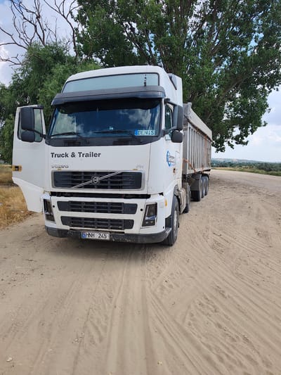 A white Volvo truck with a silver trailer is parked on a dirt road. The truck's driver's side door is open. The truck has the words "Truck & Trailer" and "VOLVO" printed on the front. A license plate with the letters "HNH 245" is visible on the front of the truck. The background features trees and a cloudy sky. The scene appears to be located in a rural area, possibly on the outskirts of Alexandru Ioan Cuza, Moldova. There are no people visible in the image.