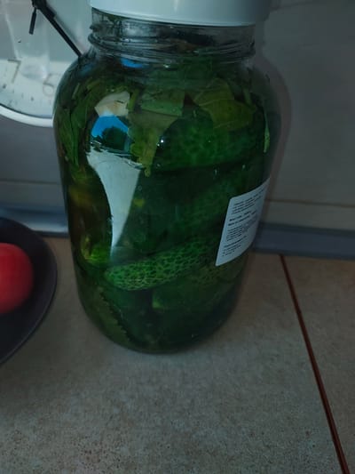 The image shows a large glass jar filled with pickled cucumbers and greenery, likely dill or other herbs, and possibly garlic. The jar has a white lid and a label with Polish text, indicating it is a food product. Next to the jar on the left is a dark bowl containing a single red tomato. In the background, a portion of a wall clock with Roman numerals is visible. The scene appears to be set on a tiled countertop, possibly in a kitchen. The label on the jar can be partially read and appears to be a product from Poland, manufactured by "AGROFRUIT S.R.L." with an address in Brzeg, Poland. The setting does not provide specific clues to determine the exact location within the city of Stargard, Poland, but suggests a domestic or commercial food preparation environment.