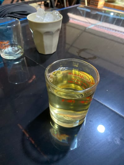 The scene depicts a tabletop arrangement of beverages and ice, situated in Đak Đoa, Vietnam. In the foreground, a clear glass contains a yellowish liquid with small bubbles near the rim; red Vietnamese text is visible through the liquid and glass. To the upper left, an empty clear glass with faint orange text rests on the same surface. A light-colored, possibly off-white or beige, faceted cup filled with ice cubes is positioned further back. All items are placed on a dark, reflective surface, likely a table, which exhibits minor scratches, dust, and reflections of the objects and ambient light. A dried, dark ring stain is visible on the table behind the full glass. The background is out of focus, showing blurred indoor elements. No individuals are visible.