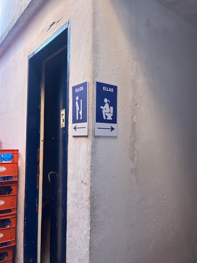 Signs in Dock Sud, Argentina indicating restrooms for men ("ELLOS" with a male figure facing right) and women ("ELLAS" with a female figure on a toilet) are affixed to a concrete wall above a doorway or opening. An additional, smaller sign is also visible on the doorway lintel. Orange and blue crates are stacked below.
