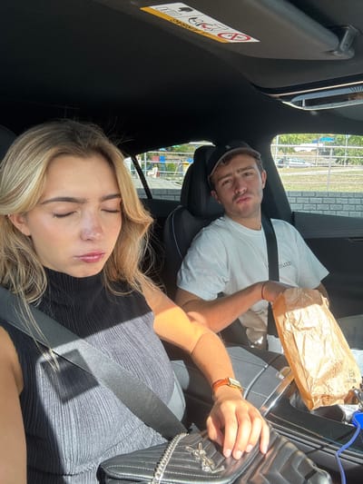 The image captures two individuals inside the dark interior of a vehicle during daylight hours, likely in an urban or suburban area of Leiria, Portugal.
**People:**
- **Female (Foreground, Left):** A young adult with blonde, shoulder-length hair, positioned in the front passenger seat. She wears a black, high-neck ribbed sleeveless top and is secured by a black seatbelt. Her eyes are closed, and her lips are slightly pursed. On her lap rests a black quilted handbag with a silver chain strap and a prominent 'YSL' logo. Her right wrist displays a rectangular-faced watch with a brown textured strap and a thin orange bracelet. Strong sunlight illuminates her right arm and the side of her face.
- **Male (Background, Right):** A young adult, seated in the driver's seat. He wears a white short-sleeved t-shirt with a dark logo and a dark baseball cap. He is secured by a black seatbelt and has a thin red bracelet on his left wrist. His mouth is puckered in a playful expression, and he holds a crumpled light brown paper bag in his left hand, resting on his lap.
**Objects and Vehicle Interior:**
- The vehicle's interior features a dark headliner with a visible "AIRBAG" warning label on the passenger-side sun visor. A sunroof is also visible.
- Dark leather seats with stitching are present.
- A modern center console is partially visible between the two individuals.
- A blue cable (possibly charging) extends from the console area towards the male's side.
**Environment (Outside the Vehicle):**
- Through the vehicle windows, an outdoor scene is visible.
- Immediately behind the female, there is a light-colored concrete or stone wall.
- Behind the male, a grey concrete wall or barrier is visible. Beyond this, a white structure adorned with distinct colorful square panels (red, blue, yellow) suggests a public space, such as a park or recreational area.
- Further in the distance, green foliage (trees) and a partial view of another white vehicle are discernible.