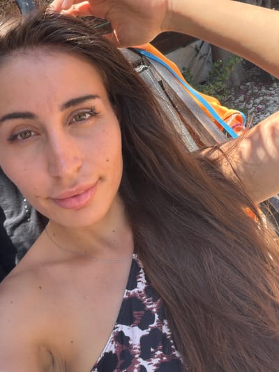 A woman with long, dark brown hair and light brown eyes is captured in a close-up selfie, taken outdoors in Mazotos, Cyprus. She is wearing a bikini top with a black and white leopard print pattern and a delicate silver chain necklace. Sunlight illuminates her face, highlighting her skin and the natural beauty of her features, including subtle freckles and a mole on her cheek. Her left hand is raised, gently touching her hair, while her right arm is visible in the background, resting on what appears to be a lounge chair made of wood and metal. The background reveals hints of a dry, rocky landscape typical of coastal Cyprus, with sparse green foliage and scattered pink flower petals on the ground. The overall scene evokes a relaxed, sunny atmosphere, possibly during a vacation or a day at the beach.