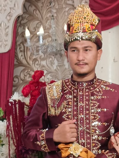 A man dressed in ornate maroon attire, including a jeweled crown, stands in what appears to be a formal setting with floral decorations and a patterned backdrop. The man has dark hair, a mustache, and is looking to his right. The attire is heavily embellished with gold thread, beads, and gemstones, with elaborate shoulder adornments. A gold sash is visible around his waist. In the background, red fabric drapes, a chandelier with electric candles, and red floral arrangements can be seen. The scene suggests a cultural or ceremonial event, possibly a wedding, within a decorated interior space in Meuliek, Indonesia.