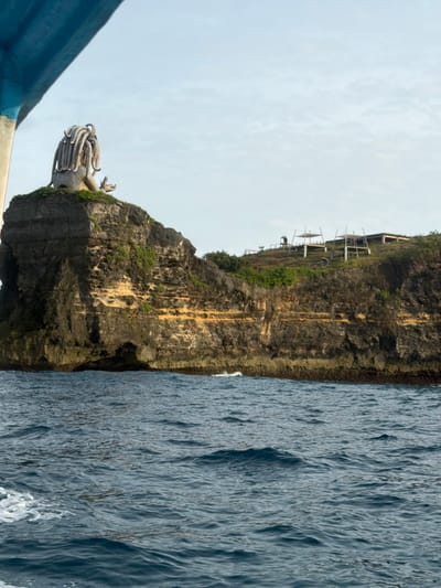 The image depicts a coastal scene in Sakti, Indonesia, viewed from the water. In the immediate foreground, the blue underside of a boat's hull or canopy partially frames the upper left corner, with white foam visible on the dark blue, rippling ocean surface. A rugged, multi-layered rock cliff dominates the midground, rising steeply from the water. Its face exhibits dark grey and brown stratifications, punctuated by prominent horizontal bands of yellowish-orange rock and sparse green vegetation. Small erosional caves are present at the waterline.

Perched atop the highest, leftmost section of the cliff is a large, light-colored statue. It portrays a stylized humanoid figure with an elaborate headpiece resembling numerous thick dreadlocks, seated or crouched. To the right of this statue, on a more gently sloping part of the clifftop, are several incomplete wooden or bamboo structures with rudimentary roofs, indicative of ongoing construction or temporary shelters. A single, small, indistinct humanoid figure is visible near these structures. The background consists of a pale blue sky with faint, scattered clouds.