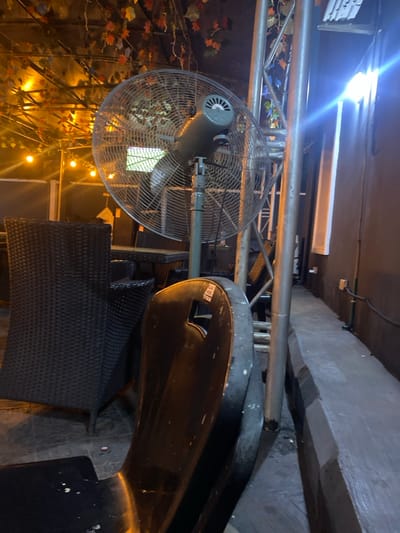 This image depicts an outdoor or semi-outdoor seating area within the city of Igbessa, Nigeria, illuminated by artificial light during what appears to be nighttime. A large, grey, industrial-style standing fan with a circular protective cage and visible rotating blades is positioned slightly left of center, indicating operation. To the left of the fan, a dark brown, tall-backed wicker chair is visible, with other similar wicker tables and chairs partially discernible in the background. In the immediate foreground, the scuffed, dark-colored backrest of a chair, possibly plastic or metal, featuring a small rectangular cutout, is angled towards the right. Overhead, a structure is draped with artificial foliage in red, orange, and green hues, interspersed with warm-toned string lights. On the right side of the frame, a bright, cool-toned rectangular light fixture is mounted on a dark wall, casting significant glare. A narrow, grey paved pathway or low bench runs parallel to this wall, separated from the main seating area by a prominent grey metal pole and other structural truss elements. No individuals are present in the scene.