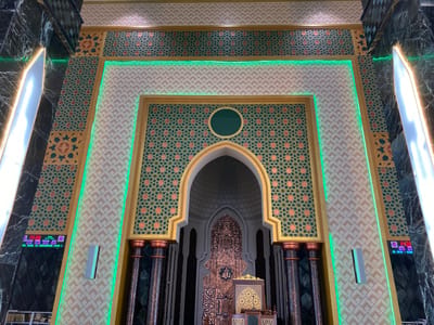 The image shows an ornate interior of a mosque in Aluekambuk, Indonesia. The central focus is a large, arched doorway leading to what appears to be the mihrab area. This archway is framed by intricate geometric patterns in shades of green, gold, and coral, highlighted by green LED lighting. Tall, dark columns with copper-colored capitals flank the entrance. Behind the arch, a richly decorated, metallic structure, possibly a pulpit or part of the mihrab, is visible, featuring Islamic calligraphy. The walls are adorned with detailed tilework exhibiting repeating geometric motifs and starburst designs. To the left and right of the central structure, digital displays show prayer times and the date. The floor is not visible, but the overall impression is one of grandeur and elaborate Islamic architectural design.
