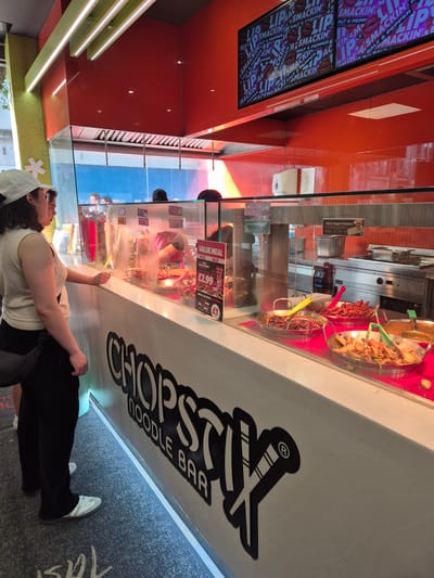 This indoor shot captures a customer at a Chopstix Noodle Bar. The location is likely in Cardiff, United Kingdom, given the architectural and environmental cues. The customer, a woman, stands at the counter, looking at the food display. The Chopstix logo is prominently featured on the counter. A menu board overhead advertises "LIP SMACKIN" meals, alongside a separate Salt & Pepper Potatoes promotion. A "VALUE MEAL" deal is advertised at £2.99. The interior is well-lit with modern lighting fixtures. Through the glass counter, we can see various food options. The environment suggests a fast-casual dining establishment.