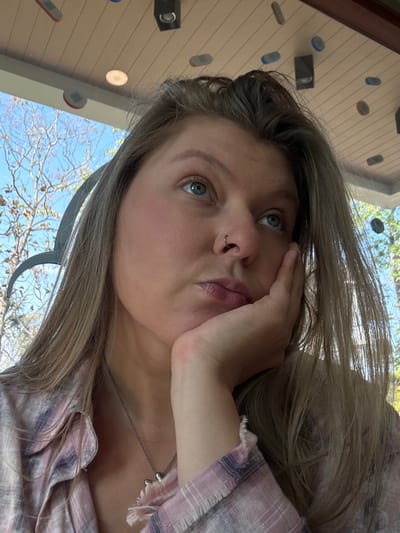 An overhead-angle portrait captures a single female subject in what appears to be a semi-outdoor or open-air structure in San Phra Net, Thailand. The subject, positioned centrally, has fair skin, long light brown hair, and blue eyes. She wears a small, circular septum piercing and a light pink and white plaid shirt with frayed cuffs, along with a thin silver necklace featuring a small pendant. Her right hand supports her chin, with her fingers lightly curled against her cheek. Her gaze is directed upwards and slightly to the right, lending a contemplative or pensive expression. The visible background consists of a light-colored slatted or paneled ceiling with multiple recessed circular lights, black rectangular fixtures resembling speakers or lights, and various small, flat, multi-colored (red, blue, grey, orange) oval and rectangular objects attached to the ceiling. Beyond this structure, a clear blue sky with some bare tree branches and sparse foliage is visible, indicating an outdoor setting during daylight hours.