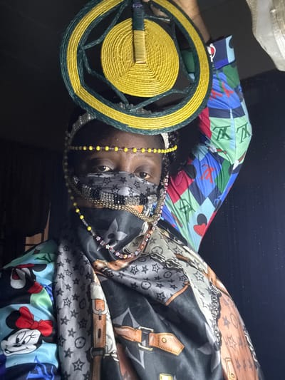 A person is lying down, facing the camera, wearing various layers of patterned clothing and beaded jewelry. The clothing includes items with Louis Vuitton logos, Mickey Mouse prints, and belt buckle designs. The person has a yellow and green coiled circular object near their head. The background is dark and blurry, obscuring details of the environment. No specific location within the city of Madangala, Nigeria, is discernible from the image.