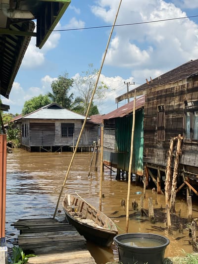 This image depicts a waterfront scene in Berangas Timur, Indonesia, characterized by wooden stilt houses lining a muddy brown river under a bright blue sky with scattered white clouds. A small wooden boat is moored to a weathered wooden jetty. The stilt houses, constructed from aged wood, exhibit signs of wear and tear, with some featuring corrugated metal roofs and others covered in a green tarp. Vertical bamboo poles are strategically placed in the water, likely serving as supports or mooring aids. The water itself appears murky and contains some floating debris. The overall impression is one of a rustic, possibly impoverished, community integrated with its riverine environment.