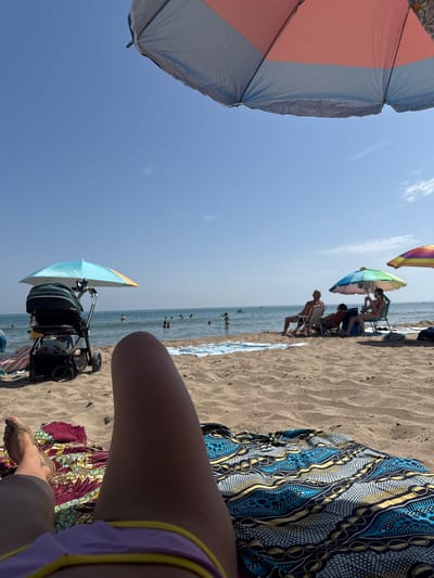 The image depicts a beach scene in Gavà, Spain, on a sunny day. In the foreground, a person's legs are visible, lying on a colorful beach towel spread on the sand. To the left, a baby stroller with a parasol shading it is parked on the sand. In the middle ground, several people are relaxing under beach umbrellas, with some sitting on chairs and others near the water's edge. The sea is calm with a few individuals swimming or wading. The background consists of the clear blue sky and the distant horizon. The environment is a typical beach setting with sand, water, and beach accessories.