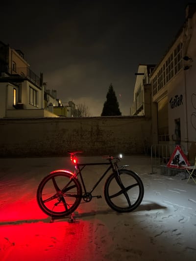 A black bicycle leans against a wall, illuminated by a red light emitting from its rear. The bike's wheels have an unusual spoke pattern. The wall is next to a narrow street, lined with buildings and houses. Above, a dark sky with a few visible stars and a tall, dark tree silhouette looms. A traffic sign stands near the wall. The image is rotated to the side.