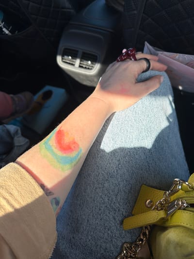 The image shows a person's arm with a colorful tattoo of layered curves resembling a rainbow. They are wearing denim jeans and a light-colored long-sleeved shirt. On their hand, they are wearing two rings, one dark and one a glossy red. The person is sitting in what appears to be the back seat of a car, with car seat fabric and air vents visible. A small, lime-green purse with a chain strap is resting on their lap. The lighting suggests it is daytime, with sunlight casting shadows. The provided location, Пяршайскі сельскі Савет, Belarus, is a rural administrative region and does not provide specific details about the scene itself, which appears to be within a vehicle.
