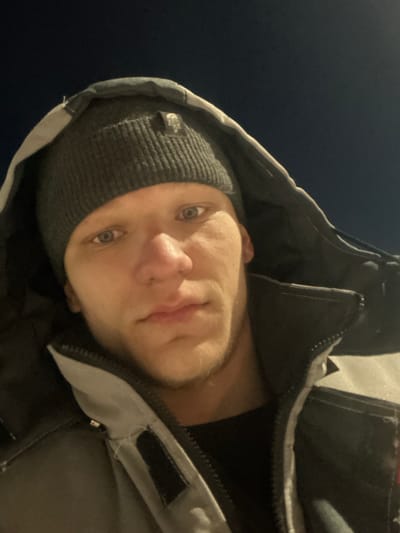 A close-up, upward-angle selfie depicts a single male individual outdoors at night. The man has light skin, blue eyes, and light stubble on his chin and jawline. He is wearing a dark grey knit beanie with a visible brand logo (appearing to be The North Face) on the front-left side, beneath the raised hood of a grey and black winter jacket. A black garment is visible under the open jacket collar. His expression is neutral as he looks directly into the camera. The background is a uniform dark blue, indicating nighttime conditions, with no discernible features, landmarks, or specific environmental details that would allow for identification of a location within the city of Залесная, Russia, or any other specific geographic area.
