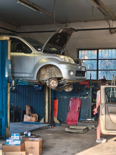 In a car repair shop in Chiugiana, Italy, a silver car is elevated on a hydraulic lift with its hood open, revealing internal engine components and showing that the front wheel has been removed. The scene is illuminated by artificial light from overhead fluorescent fixtures and natural light filtering through a large, multi-paned window in the background, suggesting it is daytime.

A young woman with dark hair is standing to the right of the car, wearing a patterned top and shorts. She is looking towards the car, her hands clasped loosely in front of her. Her posture and expression do not convey a strong emotion, suggesting a neutral or observant demeanor.

The shop floor is concrete and appears somewhat cluttered with various automotive parts and tools. Visible objects include several cardboard boxes, some stacked on top of others, a spare tire, coil springs, a red plastic tool tray, and various tools hanging on a blue metal wall. A sign with Italian text, "ATTENZIONE: MUFARE," is visible on the wall. The overall setting suggests a working environment dedicated to vehicle maintenance and repair. The presence of the woman and the partially disassembled car indicate an ongoing repair job. The light suggests it could be late afternoon or early morning, given the warm tones coming through the window.