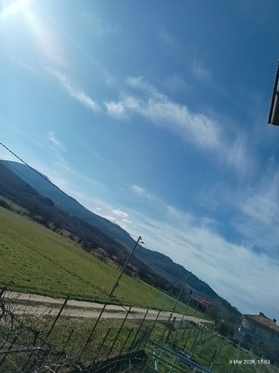 The image displays a rural landscape in Voiniagovo, Bulgaria, captured on a sunny afternoon at 15:03 on March 9, 2026. A bright blue sky with elongated wispy cirrus clouds covers the upper portion of the frame, with the sun visible as a bright glare in the upper left quadrant. A prominent mountain range, partially covered with bare deciduous trees, dominates the background, sloping from left to right. In the midground, a wide green field stretches to the left of an unpaved dirt path that horizontally traverses the scene. Several utility poles with overhead lines run alongside this path, extending into the distance. On the far right of the midground, a building with a reddish-brown tiled roof and light-colored walls is visible. The immediate foreground consists of bare, tangled branches, likely from a bush or tree, and a metal wire fence. Additional metal structures, possibly agricultural in nature, are present behind the fence on the right. No individuals are present in the scene.