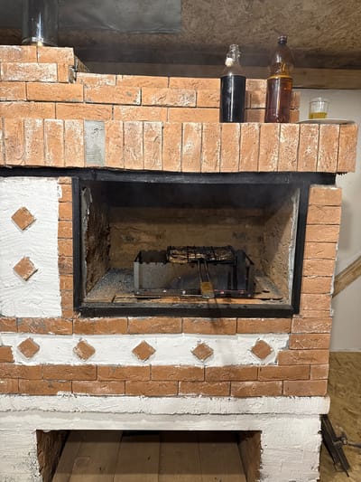 The image shows a brick barbecue oven located in Gon'ba, Russia. The oven has an open firebox containing a metal grill with food. On top of the oven, there are two bottles, one dark and one amber, and a glass with a yellow liquid. The oven is made of red bricks with white decorative elements and is situated indoors, with wooden flooring and ceiling visible. The overall scene suggests a setting for cooking and dining.