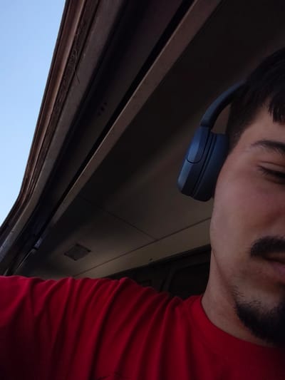 A close-up, waist-up, eye-level, outdoor, daytime, low-angle shot captures a young man wearing a red t-shirt and blue headphones. He is positioned on the right side of the frame, facing left, with his head tilted slightly down and his eyes closed. The background consists of the interior of what appears to be a train carriage, with a window visible at the top left showing a clear blue sky. The image is dark, with the man's face and t-shirt illuminated by natural light coming from the window. The overall atmosphere is peaceful and contemplative. The location is not specified in the image.