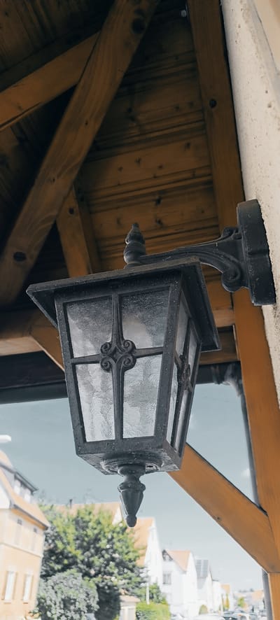 An outdoor wrought iron lantern hangs from a dark metal bracket attached to a textured, light-colored building exterior. The lantern has a rectangular glass pane design with decorative metal crossbars. Above the lantern, a wooden roof structure with exposed beams is visible. In the background, out of focus, are residential buildings with light-colored facades and orange-tiled roofs, as well as green trees and a bright sky. The overall scene appears to be an exterior shot of a building in Schwaikheim, Germany, likely during daytime.
