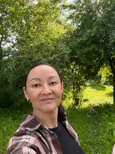 A woman with dark hair and brown eyes takes a selfie outdoors in a natural setting. She is wearing a pink and brown plaid shirt over a black top. The background consists of lush green trees and grass, with sunlight filtering through the leaves, creating a dappled effect. The location is the village of Bolshoy Turysh, Russia.