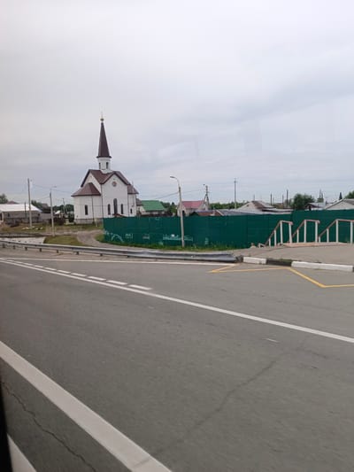In Zudilovo, Russia, a white church with a brown, pointed steeple topped with a cross stands to the left of a bright green metal fence. The church has a brown roof and two rows of windows. The fence runs horizontally across the middle of the image, separating the church and surrounding buildings from a grey asphalt road in the foreground. White dashed lines mark the lanes on the road, and a white curb with yellow markings is visible. Beyond the fence, other buildings with varied roof colors and styles are scattered. The sky is overcast and grey.