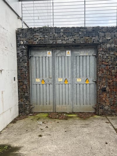 The image shows a pair of industrial metal doors set within a gabion wall structure. The doors are grey and ribbed, with two "Danger Keep Away" warning stickers on the left door and two similar stickers on the right door, one of which also displays a lightning bolt symbol. Above the right door, text reads "FINGAL BAY B PK 065305Z". The gabion walls are constructed from wire cages filled with grey and reddish-brown stones, flanking the doors. To the left of the gabion wall is a textured, weathered white concrete wall. The ground in front of the doors is a grey concrete surface with some scattered debris including leaves, twigs, and a crumpled plastic bottle. The scene appears to be outdoors, possibly in an industrial or service area, with a cloudy sky visible through a metal grid structure at the top of the frame. The presence of the "FINGAL BAY B PK" marking suggests a location within or related to Fingal Bay, which is in Balbriggan, Ireland.