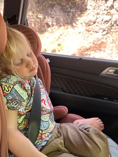 A child with blond hair, wearing a colorful shirt with "GUESS" and "LOS ANGELES" printed on it, is sleeping in a car seat. The child is secured with a seatbelt. Their bare feet are visible next to tan-colored pants. The car's interior includes black leather door panels. The window shows a blurred view of outdoor foliage. The location is not in Ula, Turkey.