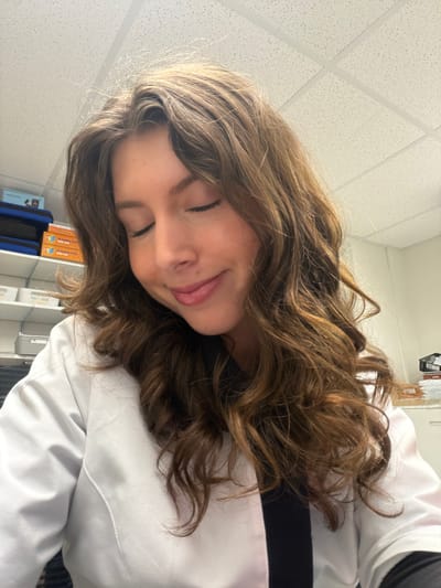 A young woman with long, wavy brown hair is lying down, with her eyes closed and a slight smile. She is wearing a white lab coat over a black shirt. The background suggests a sterile environment, possibly a lab or classroom, with a white ceiling and what appears to be shelving containing books or folders. The location is unknown, but the request specifies Longueuil, Canada.
