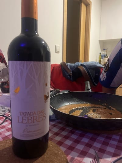 The scene depicts an indoor setting, identified as a residential space in Pedrouços, Portugal. In the foreground, a dark glass bottle of "TAPADA DAS LEBRES Premium" red wine is positioned on a cork coaster, resting on a red and white checkered tablecloth. The wine bottle features a white label with an illustration of light-colored tree silhouettes and two small rabbit-like figures, one golden and one brown. To the right of the wine bottle, a black frying pan contains a small quantity of viscous, brownish-orange liquid, likely a sauce, with a small metallic utensil resembling a slotted spoon or strainer scoop resting within it. A silver fork is partially visible on the tablecloth in the bottom right corner. Dark cables or wires are visible on the left side of the tablecloth.
In the midground, a person's upper torso and arm are partially visible to the right, wearing a blue and white striped shirt, consistent with a sports jersey. Behind the objects on the table, a red fabric item and a blue denim-like item are present. The background consists of a light-colored wall with a prominent light brown wooden door frame and a white light switch. Further to the right in the background, a section of a kitchen area with white cabinets or shelving is visible, displaying several bottles, including what appears to be olive oil and water. The lighting in the scene is artificial, with a noticeable purple tint present.