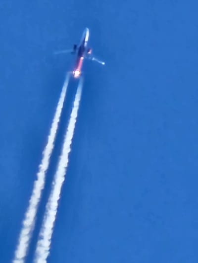 A white and red airplane soars across a clear blue sky, leaving behind two thick white contrails. The plane is viewed from behind and below, with a vibrant burst of orange flame emanating from its engine. The contrails stretch out horizontally, gradually fading as they extend into the distance. The overall scene conveys a sense of speed and altitude.