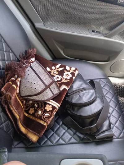 In Yangikurgan, Uzbekistan, inside a car, a brown prayer mat with floral patterns in white and gold is laid out on a quilted black leather car seat. A black leather sling bag with a zipper closure rests on top of the prayer mat, its strap draped across it. The car's interior door panel, made of grey and textured plastic, is visible in the upper portion of the image, with a speaker grille on the right. The scene appears to be set within the passenger area of the vehicle.